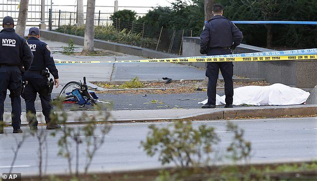 A New York City police officer stands next to a body covered in a white sheet near a vandalized bicycle along a bike path.