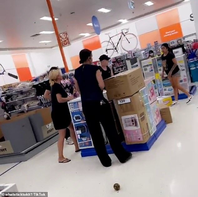 A woman with bleached blonde hair confronted the dark-haired girl as they stood around stacked products for sale with two men, while a tall staff member looked on.