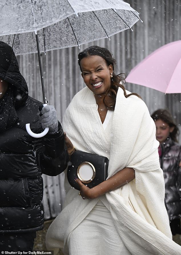 A woman wearing an all-white outfit trekked through the snow as someone held an umbrella over her to attend the fashion show