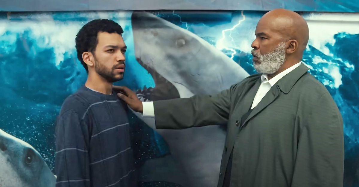 An older man with a beard places his hand on the shoulder of a younger man in front of a shark poster.