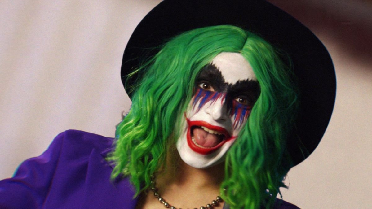 Vera Drew as Joker the Harlequin, a mashup of Joker and Harley Quinn, in green wig, clown face paint with a big red lipstick smile, a purple suit jacket and black hat in The People's Joker