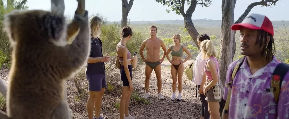 A group on a walk in Australia.  In the foreground a black man stares intently at a koala.  Meanwhile, a handsome blonde man wearing only panties and a beautiful blonde woman stand with their arms crossed in the background.  Everyone else in the group gapes at them.