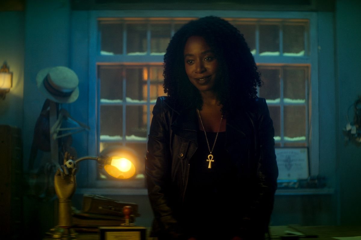 Kirby Howell-Baptiste as Death in Dead Boy Detectives.  She is dressed all in black, with a gold ankh necklace, and stands in a blue-hued room.