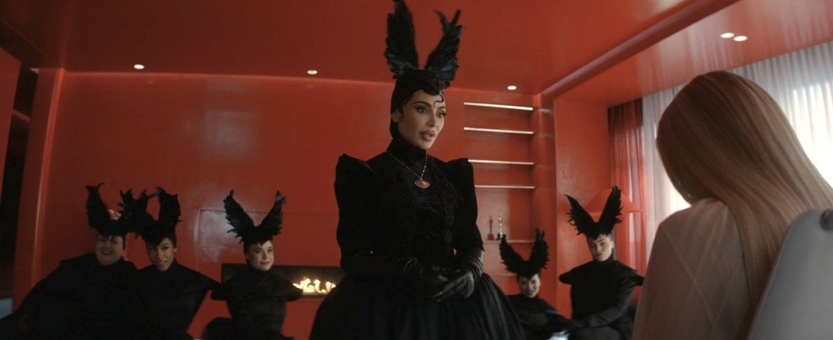 Kim Kardashian in a flashy black feather headdress leers over Emma Roberts.  Behind her is a group of women wearing the same headdress.  They are all in a bright red modern loft. 