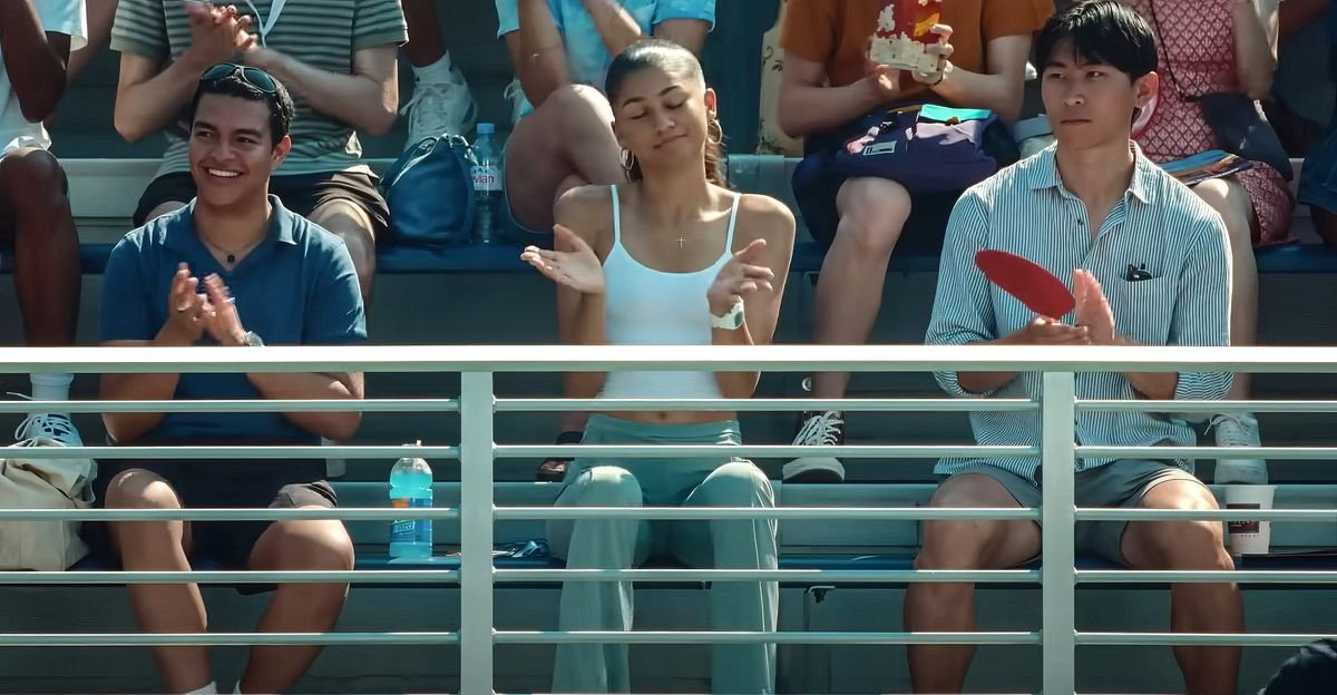 Tennis player Tashi (Zendaya) sits in the stands during a match in Luca Guadagnino's Challengers.  The fans around her applaud something happening on the field, but she smiles and shrugs, eyes closed.
